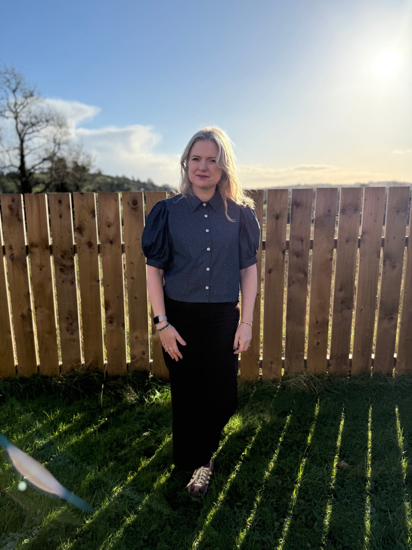 Woman standing outdoors in front of a wooden fence with grass and sky in the background