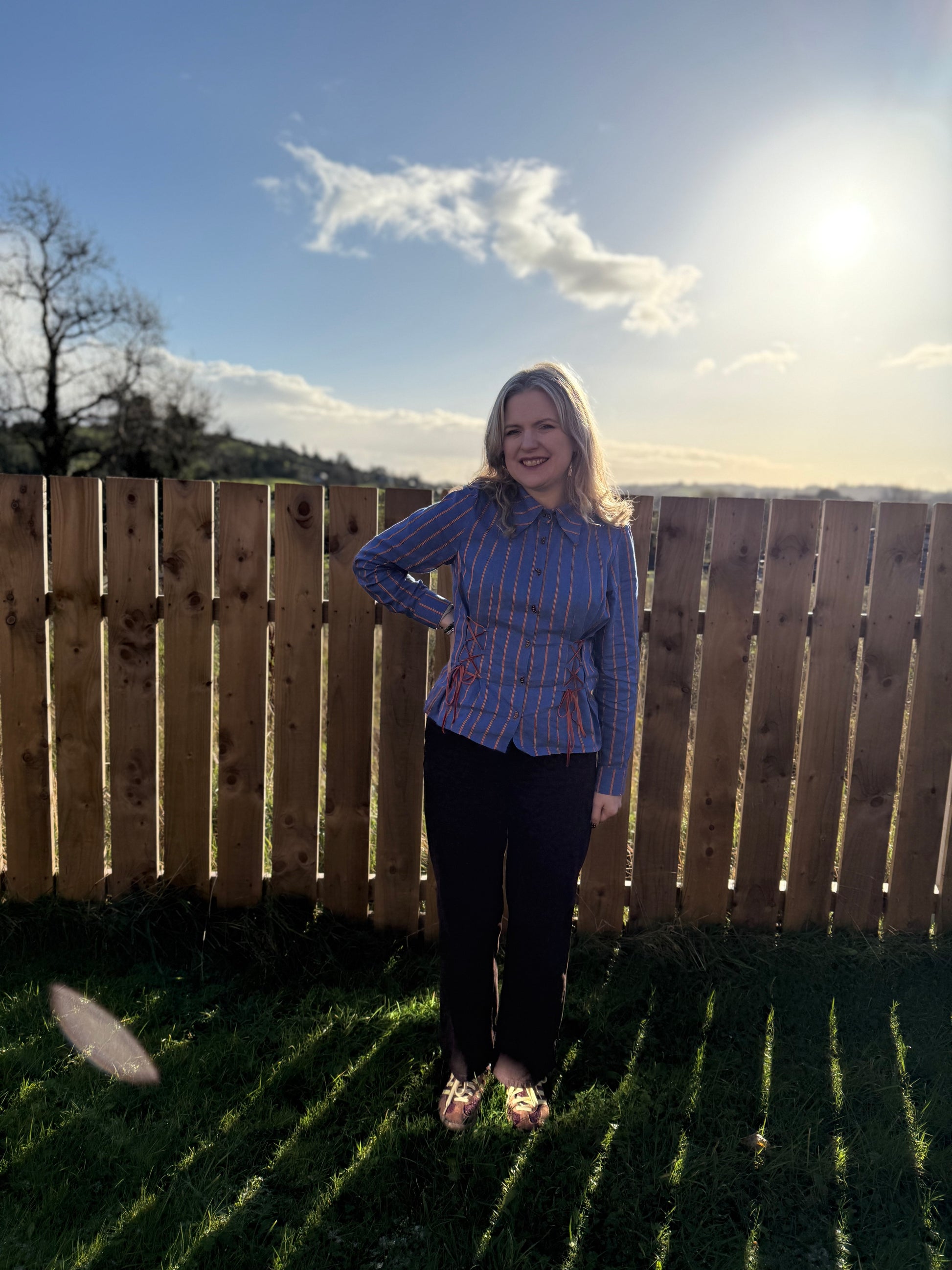 Woman wearing and blue stripe shirt, standing in a backyard with a wooden fence and clear sky.