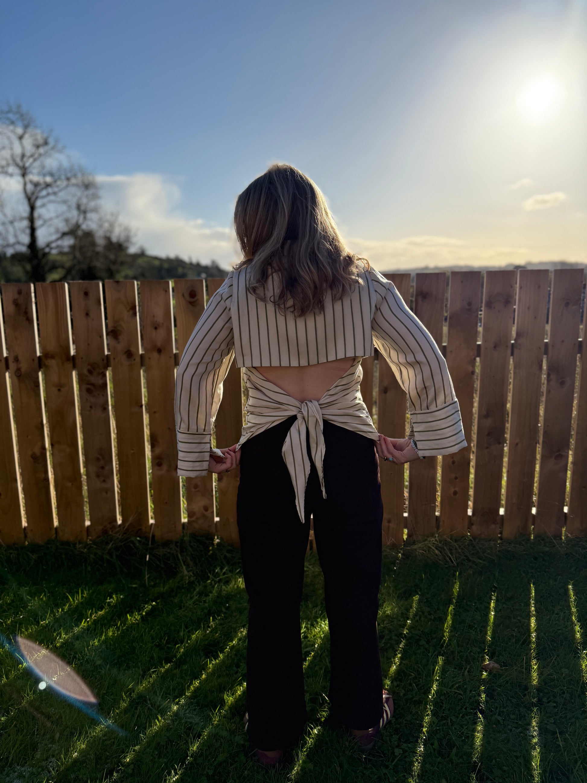 Person standing in a backyard with a wooden fence and grass, wearing a striped shirt and dark pants.