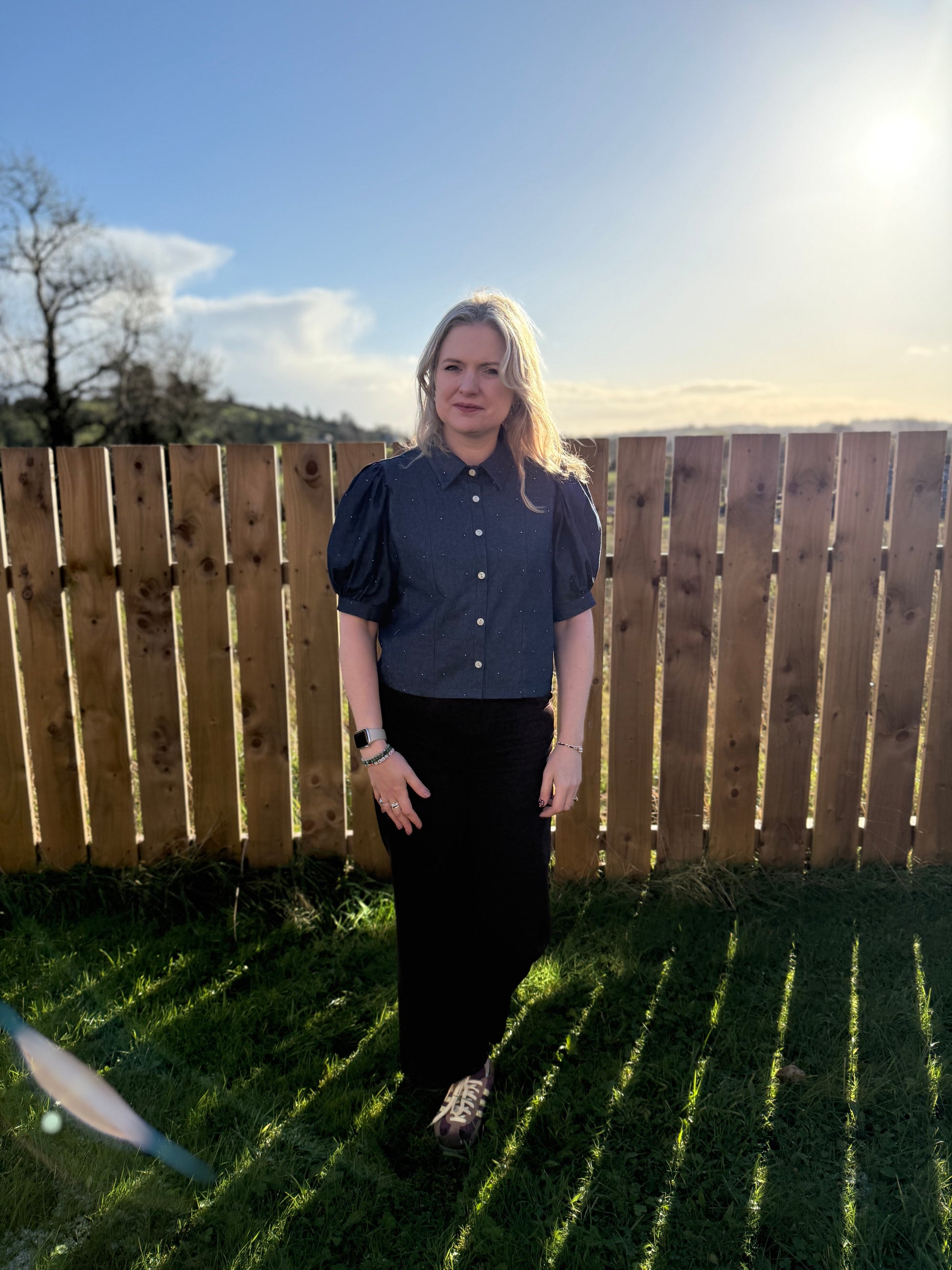 Woman standing outdoors in front of a wooden fence with grass and sky in the background